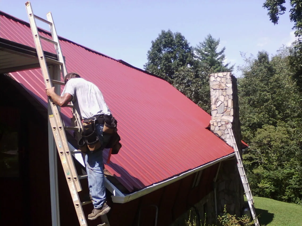 Roofer on a ladder beside a red metal roof and stone chimney — Soffit & Fascia Repair by Roofing Brothers in East Montpelier